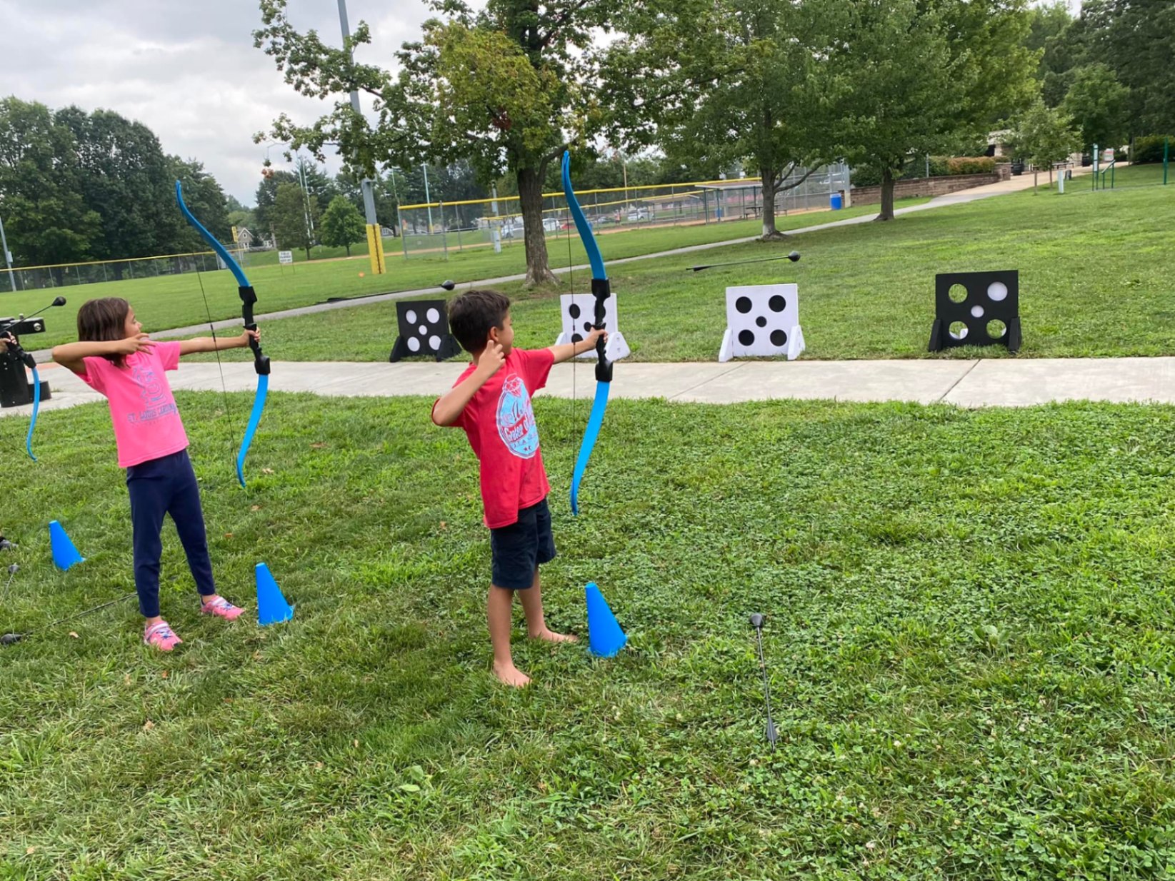 Two kids drawing foam-tipped bows at outdoor archery targets